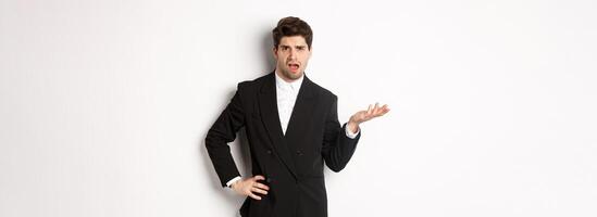Portrait of arrogant man in black suit, looking confused and disappointed, complaining about something strange, standing over white background photo