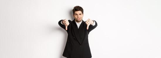 Portrait of skeptical and disappointed man in black suit, frowning upset, showing thumbs-down, dislike something bad, standing over white background photo
