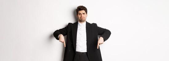Portrait of skeptical and unamused bearded man, pointing fingers down with disappointed face, standing in suit and complaining photo