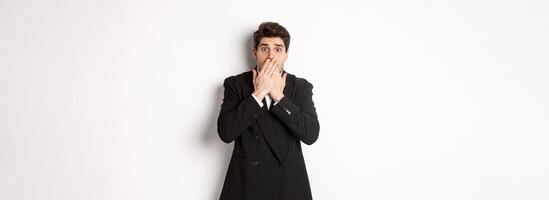Scared man in formal suit, gasping and looking frightened at camera, standing against white background photo