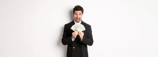 Image of handsome bearded guy in black suit, looking at money with excitement, standing over white background photo