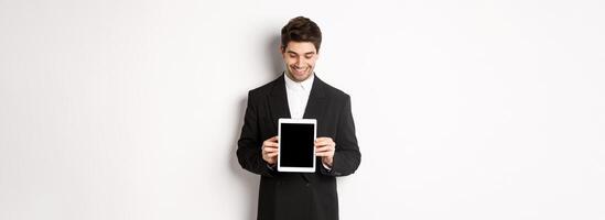 Image of good-looking male entrepreneur in black suit, looking down at digital tablet screen and showing advertisement, standing against white background photo