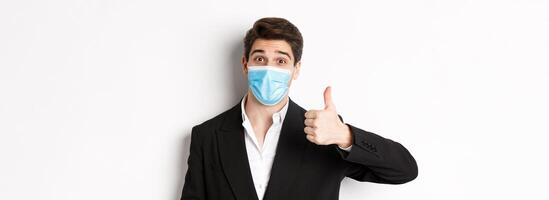 Concept of covid-19, business and social distancing. Close-up of happy businessman in black suit and medical mask, showing thumbs-up, making a compliment, white background photo
