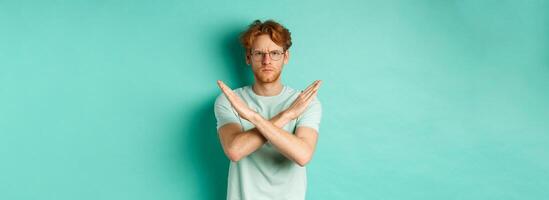 Serious and confident redhead man in t-shirt and glasses saying no, showing cross gesture to stop you, refucing or declining something, standing over turquoise background photo