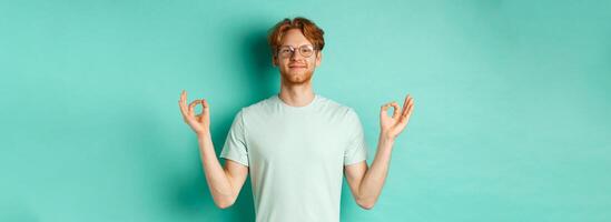 Calm and relaxed redhead man in glasses smiling, holding hands spread sideways with om mudra gesture, meditating peaceful, standing over mint background photo