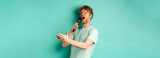 Passionate redhead man in t-shirt singing serenade with microphone, looking aside at karaoke and gesturing, standing over mint background photo