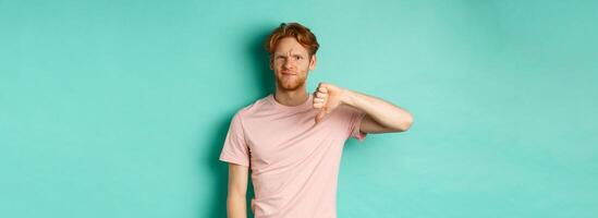 Disappointed redhead man showing thumbs-down, frowning and looking skeptical, epxress dislike and disapproval, standing over turquoise background photo