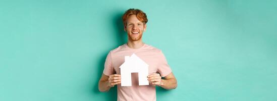 Real estate concept. Young man with red hair, wearing t-shirt, showing paper house cutout and smiling happy, standing over mint background photo