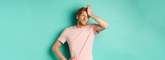 Annoyed and bothered redhead male model showing facepalm and roll eyes, standing over mint background photo