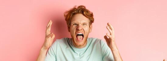 Face of angry redhead man shouting and shaking hands furious, staring outraged and cursing, express hatred and aggression, standing frustrated over pink background photo