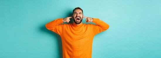 Frustrated and annoyed man shouting at neighbours upstairs, shut ears with fingers, loud bothering noise, standing over light blue background photo