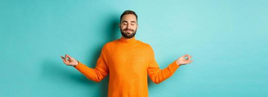 Image of relaxed and relieved man close eyes and smiling, feeling stress-free, meditating with calm expression, standing over light blue background photo