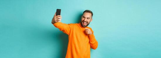 Handsome man calling on mobile phone, taking selfie and pointing at smartphone camera, standing over turquoise background photo