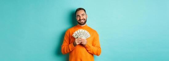 Man thinking about shopping, holding money and smiling satisfied, looking at upper left corner, standing over light blue background photo
