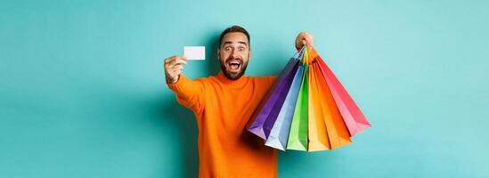 Happy aduly man showing credit card and shopping bags, standing against turquoise background photo