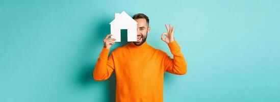 Real estate concept. Satisfied man recommending agency, showing okay sign and house maket, standing over blue background photo