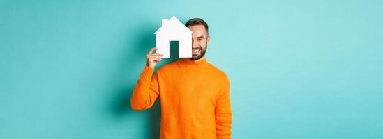 Real estate concept. Happy young man searching for home rent, holding house paper maket and smiling, standing over blue background photo