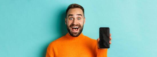 Close-up of handsome bearded guy in orange sweater, showing smartphone screen and smiling, showing promo online, turquoise background photo
