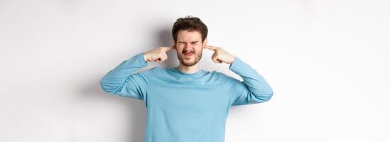 Annoyed young man plug ears from loud awful sound, being disturbed by bothering noise, grimacing displeased, standing on white background photo