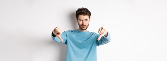 Skeptical handsome guy with beard, showing thumbs down in disapproval, dislike something bad, standing over white background photo