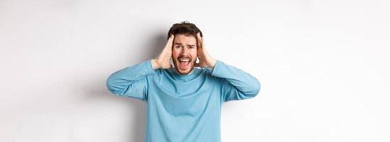 Image of young caucasian man screaming in panic, looking frustrated and shouting at camera, standing on white background photo