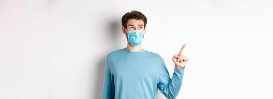 Coronavirus, health and quarantine concept. Curious guy in medical mask, pointing and looking at upper left corner banner, standing on white studio background photo