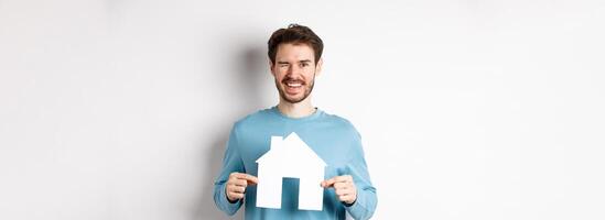 Real estate and insurance concept. Happy young man winking and smiling, showing paper house cutout, standing over white background photo