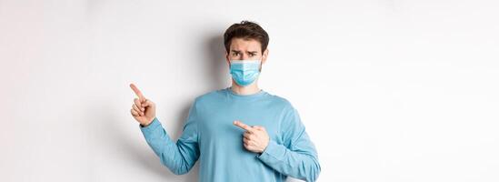 Coronavirus, health and quarantine concept. Sad man in medical mask frowning, pointing fingers right with doubtful face, standing over white background photo
