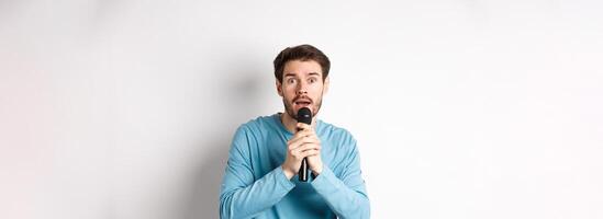 Confused young man looking nervously at camera while singing karaoke, holding microphone, standing over white background photo