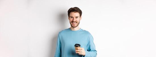 Cheerful man drinking coffee from cafe take-away and smiling, standing with paper cup in white background photo