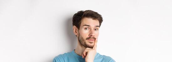 Image of thoughtful young man with beard, making choice, touching lip and looking pensive at camera, deciding something, white background photo
