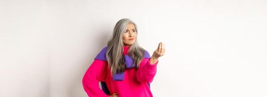 Grumpy asian grandmother complaining, showing italian what do you want gesture, shaking fingers and staring angry left at copy space, standing over white background photo