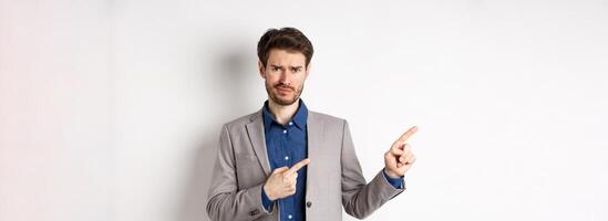 Bad idea. Displeased skeptical guy in business suit smirk and frown, pointing fingers left at upsetting news, looking hesitant at camera, white background photo