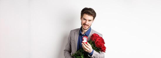 Romantic man with boquet of red roses asking to marry him, holding engagement ring and looking confident at camera, standing in suit over white background photo