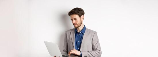 Handsome male entrepreneur working on laptop, looking serious at screen, standing against white background photo