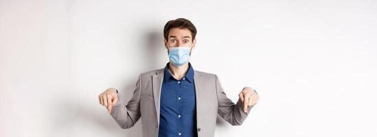 Covid-19, pandemic and business concept. Surprised male ceo manager in medical mask and suit pointing fingers down, looking amused, white background photo