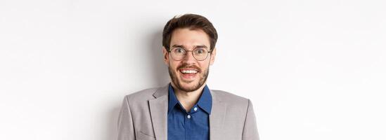 Cheerful european man in glasses and suit smiling excited, looking at camera with amazed happy face, white background photo