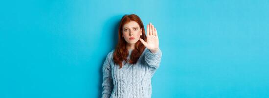 Serious and confident redhead girl telling to stop, saying no, showing extended palm to prohibit action, standing over blue background photo