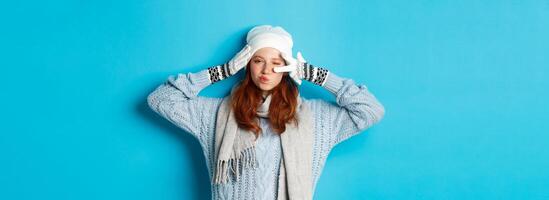 Winter and holidays concept. Happy redhead girl in beanie hat, scarf and gloves showing peace sign and pucker lips sassy, staring at camera, wearing sweater, posing over blue background photo