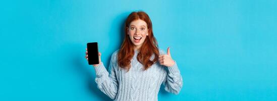 Smiling redhead girl showing smartphone screen, holding phone and demonstrating app, making thumb up in approval, recommending online store, blue background photo