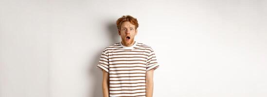 Shocked and scared redhead man screaming, open mouth and staring terrified at camera, standing over white background photo