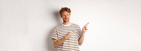 Doubtful redhead guy pointing fingers left at looking skeptical at camera, standing over white background photo