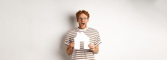 Happy and surprised redhead man winning house, holding paper home model and staring at camera, standing joyful over white background photo