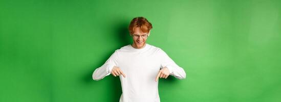 Handsome redhead man in glasses and white long-sleeve checking out advertisement, pointing fingers and looking down with pleased smile, standing over green background photo