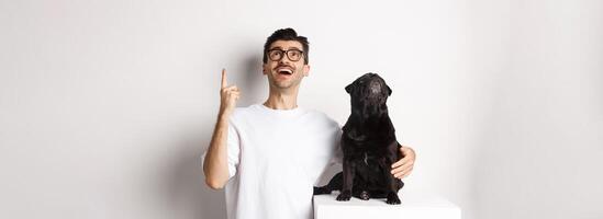 Image of happy young man and dog both looking up at promo, owner pointing finger at top, pug staring at loog, standing over white background photo