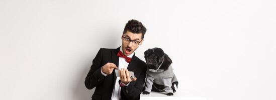 Amazed man showing to dog, pointing at mobile phone and look excited while pug sitting unbothered in costume, white background photo