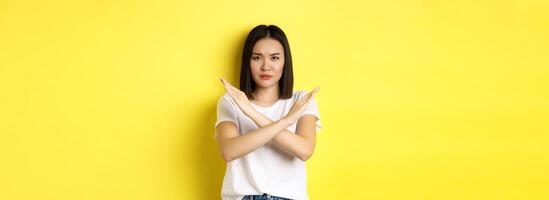 Serious and assertive korean woman showing cross stop gesture, frowning and telling no, prohibit action, disapprove something bad, standing over yellow background photo