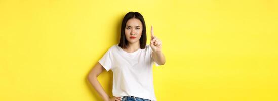 Confident and serious woman tell no, showing extended finger to stop and prohibit something bad, frowning and looking at camera self-assured, standing over yellow background photo
