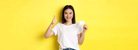 Young asian woman in casual white t-shirt showing plastic credit card and thumb up gesture, approve and recommend, smiling at camera, yellow background photo