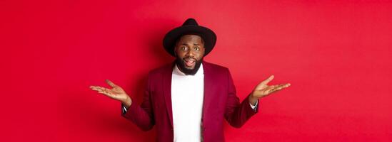 Image of confused Black man asking question, spread hands sideways and staring camera clueless, standing against red background photo